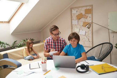 Baba ve oğul birlikte okul ödevi yapıyorlar. Küçük kız kardeşi arka planda kanepede oynarken çocuk dizüstü bilgisayar kullanıyor ve öğreniyor.