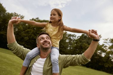 Küçük heyecanlı kız parkta babasıyla oynuyor, omuzlarında oturuyor ve gülümsüyor, birlikte harika vakit geçiriyorlar ve eğleniyorlar.
