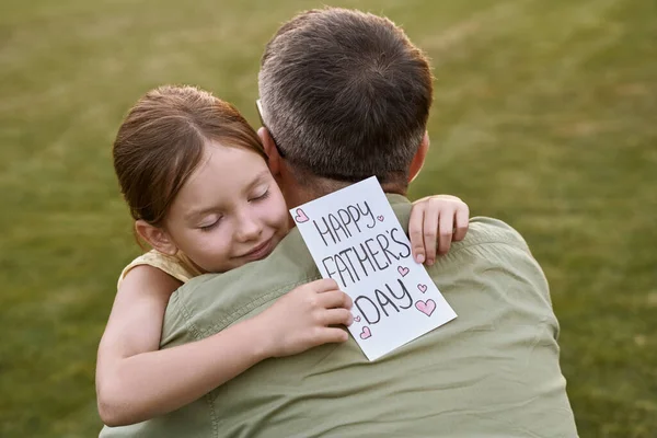 Feliz Día del Padre. Linda niña, pequeña hija abrazando a su padre y dándole postal hecha a mano mientras pasan tiempo juntos al aire libre — Foto de Stock