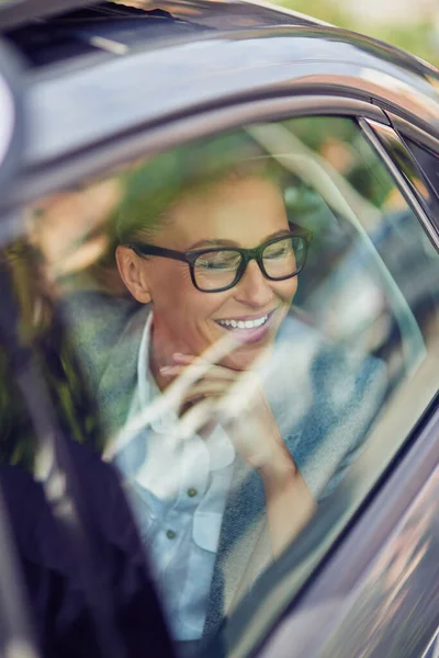 Perjalanan untuk bekerja. Wanita bisnis paruh baya yang cantik dan bahagia mengenakan kacamata yang melihat keluar dari jendela mobil dan tersenyum sambil duduk di kursi belakang dengan taksi — Stok Foto
