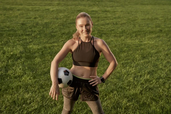 Jugador de fútbol. Hermosa mujer feliz sosteniendo la pelota de fútbol y sonriendo a la cámara mientras está de pie al aire libre en un campo verde. Jugar al fútbol —  Fotos de Stock