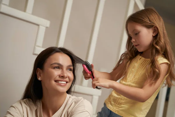 Mujer joven, feliz madre encantadora mirando a un lado y sonriendo mientras su pequeña hija peinando el pelo — Foto de Stock