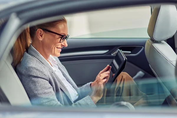 Jaringan selancar dalam perjalanan ke kantor, pemandangan indah perempuan bisnis yang bahagia duduk di kursi belakang mobil, menggunakan tablet digital dan tersenyum — Stok Foto
