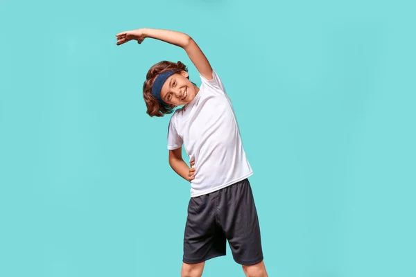 Zahřívám se. Šťastný dospívající chlapec ve sportovním oblečení dívá pryč a usmívá se při protahování jeho tělo před cvičením, stojí izolovaný nad modrým pozadím — Stock fotografie