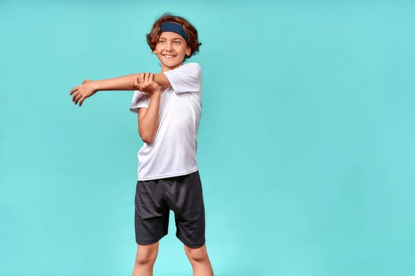 Щасливий хлопчик-підліток у спортивному одязі дивиться вбік і посміхається, розтягуючи руки перед тренуванням або тренуванням, стоїть ізольовано на синьому фоні — стокове фото