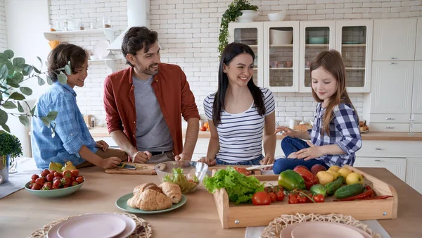 Familia vegana. Madre, padre y dos pequeños niños emocionados cocinando juntos en la cocina en casa. Preparan una ensalada, hablan y sonríen — Foto de Stock