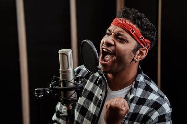 Genç bir adamın portresi, hip hop sanatçısı odaklanmış görünüyor, profesyonel bir stüdyoda şarkı kaydederken mikrofona şarkı söylüyor.