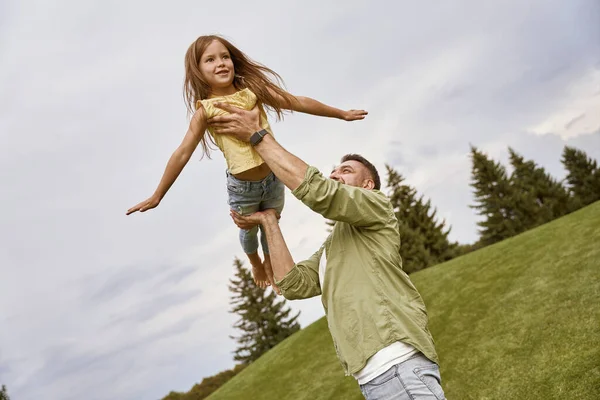 Flying with daddy. Young loving father playing and having fun his cute and happy little daughter while visiting beautiful green park on a warm summer day