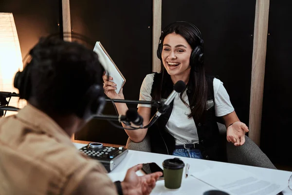 Portait of happy female radio host laughing, listening to male guest, presenter and holding a script paper while moderating a live show in studio