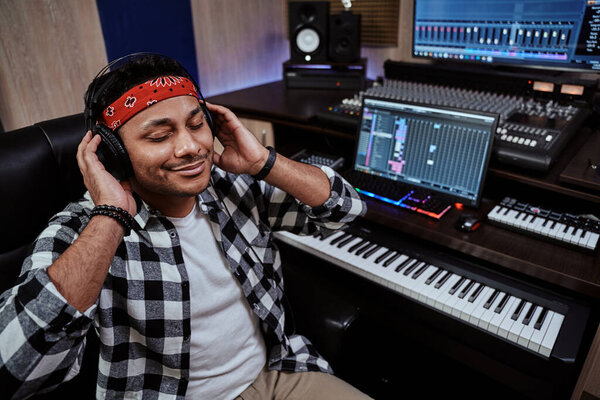 Young man, male artist in headphones sitting with eyes closed while listening to music in recording studio