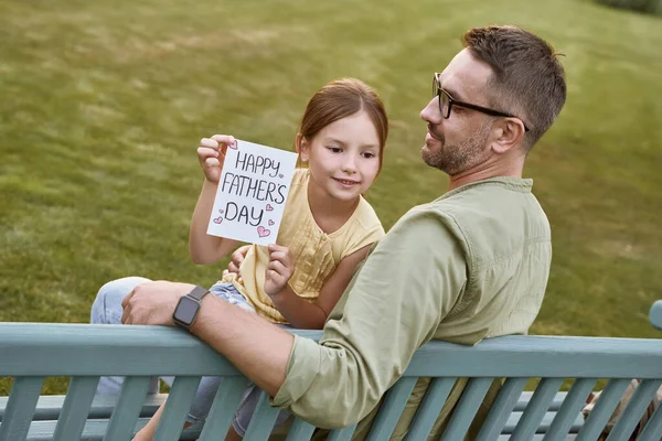 Celebrating Happy Fathers Day outdoors. Young loving father sitting on the wooden bench in park with his cute little daughter, happy girl holding handmade postcard for daddy