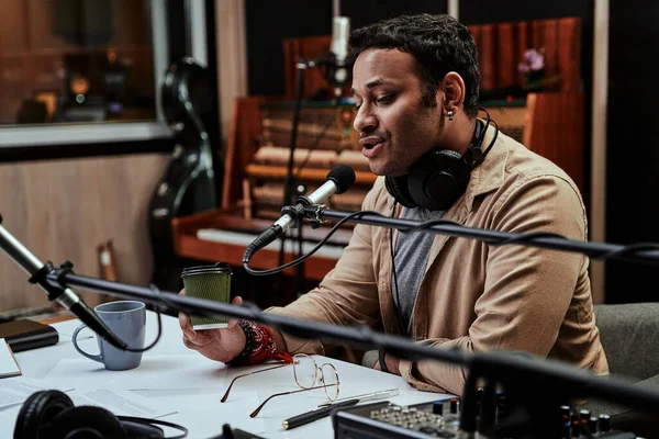 Retrato de jovem apresentador de rádio masculino tomando uma bebida, olhando focado ao falar em microfone, moderando um show ao vivo — Fotografia de Stock