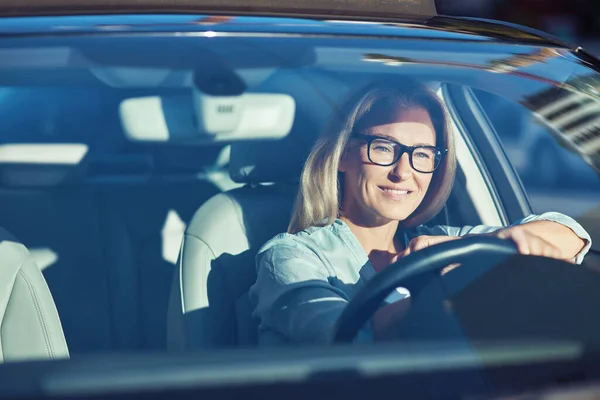 Šťastný atraktivní běloška nosí brýle řízení její moderní auto přes město na slunný den, ona se dívá na silnici a usmívá — Stock fotografie