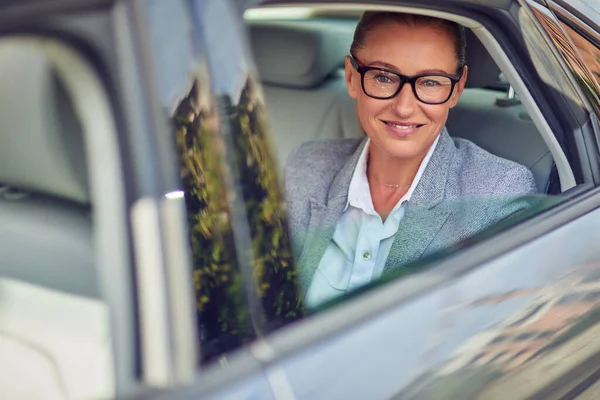 Szczęśliwy w średnim wieku kobieta biznesu w okularach patrząc przez okno samochodu podczas siedzenia na tylnym siedzeniu, ona idzie na spotkanie taksówką — Zdjęcie stockowe