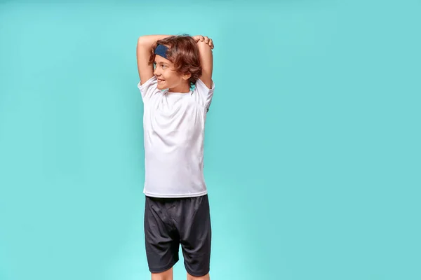 Щасливий хлопчик-підліток у спортивному одязі дивиться вбік і посміхається, розтягуючи тіло перед тренуванням, стоїть ізольовано на синьому фоні — стокове фото