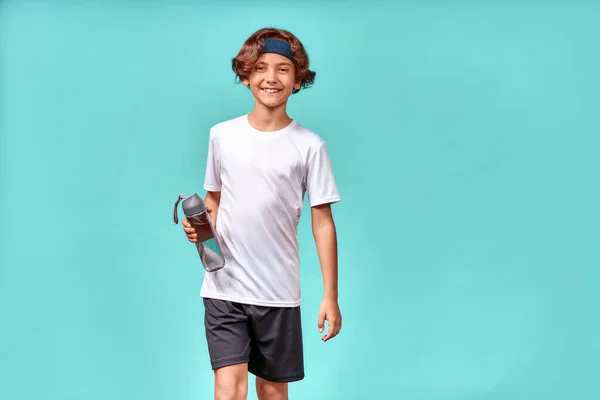 Un ragazzo carino con una bottiglia d'acqua guardando la fotocamera e sorridendo, riposandosi dopo l'allenamento o l'allenamento mentre in piedi isolato su sfondo blu — Foto Stock