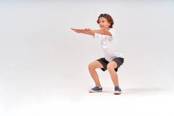 Liikuntaa. Täysi pituus valkoihoinen teini-ikäinen poika urheiluvaatteet kyykyssä, tekee vatsalihasliikkeitä eristetty yli harmaa tausta studiossa — kuvapankkivalokuva