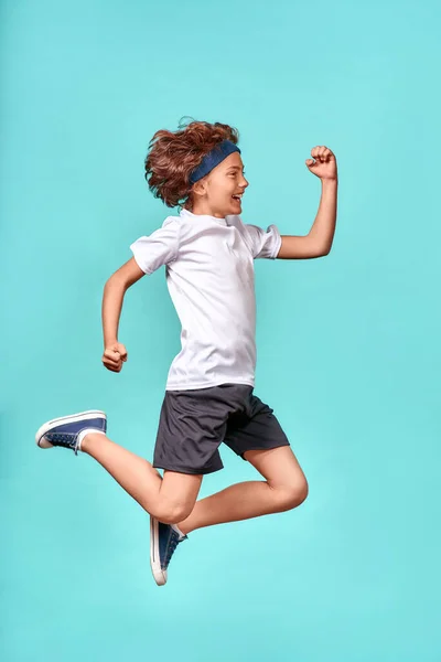 Hareket halinde. Mavi arka planda izole bir şekilde atlayan mutlu, enerji dolu genç bir çocuğun tam boy dikey çekimi.