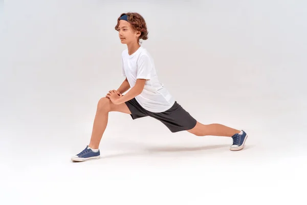 Ćwiczenia rozciągające. Pełna długość białego, sportowego nastolatka w odzieży sportowej rozgrzewającego się przed treningiem, stojącego na szarym tle w studio — Zdjęcie stockowe