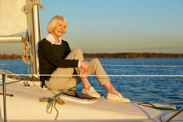 Успокойся и расслабься. Красивая пожилая женщина, сидящая на борту парусника или палубы яхты, плавающая в спокойном синем море на закате, наслаждаясь удивительным видом и улыбкой