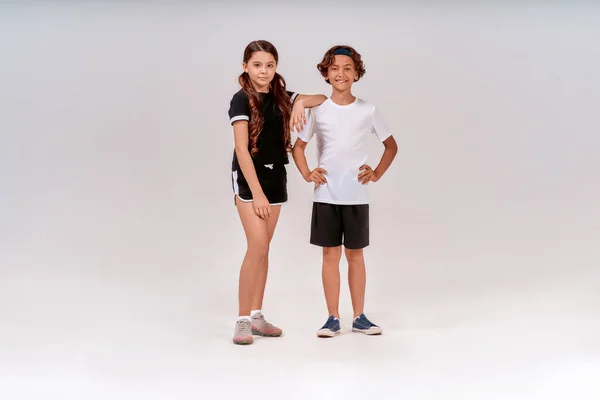 Готуємося до вправ. Двоє підлітків, милий хлопчик і дівчинка в спортивному одязі дивляться на камеру і посміхаються, позуючи ізольовано на сірому фоні — стокове фото