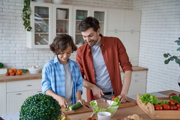 Cooking dinner. Young father teaching his son how to cut vegetables, preparing a salad while standing in the modern kitchen at home — Stock Photo, Image