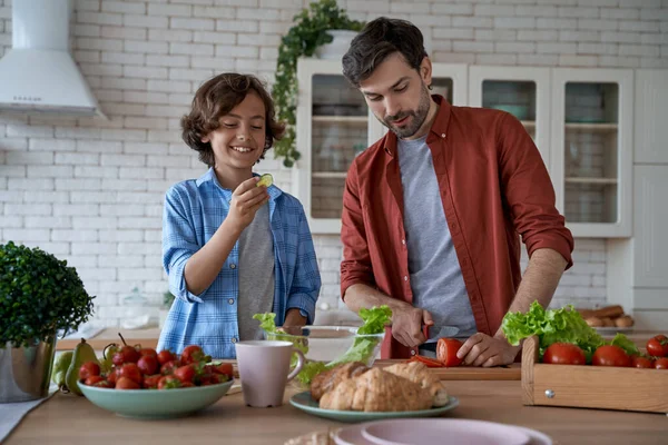 Enjoying weekend at home. Young dad and little happy son cutting fresh vegetables, preparing a salad together while standing in the modern kitchen — Stock Photo, Image