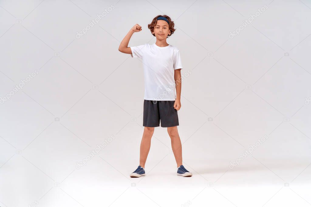 Strong kid. Full length of a strong teenage boy in sportswear looking at camera and showing his biceps while posing isolated over grey background in studio