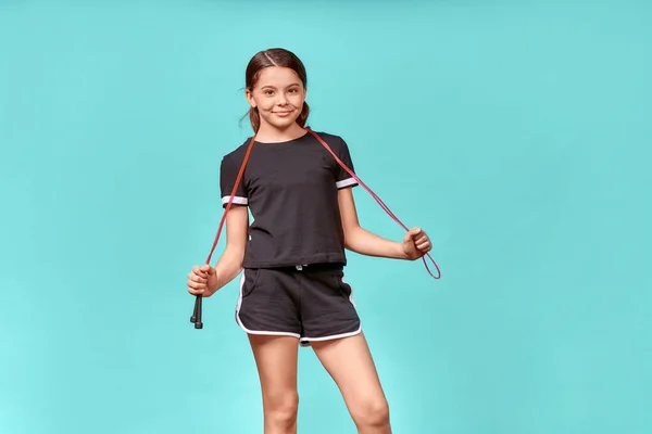 Симпатична дівчина-підліток в чорному спортивному одязі зі стрибками на мотузці дивиться на камеру і посміхається під час тренувань, стоїть ізольовано на синьому фоні — стокове фото