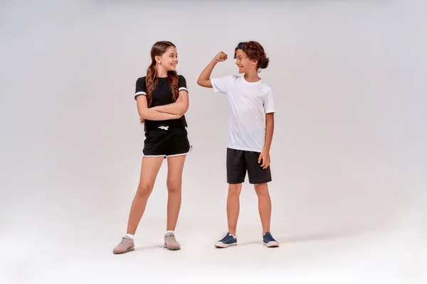 Dwoje nastolatków ćwiczących razem. Chłopiec pokazując swoje bicepsy do cute girl, stoją one odizolowane na szarym tle w studio — Zdjęcie stockowe