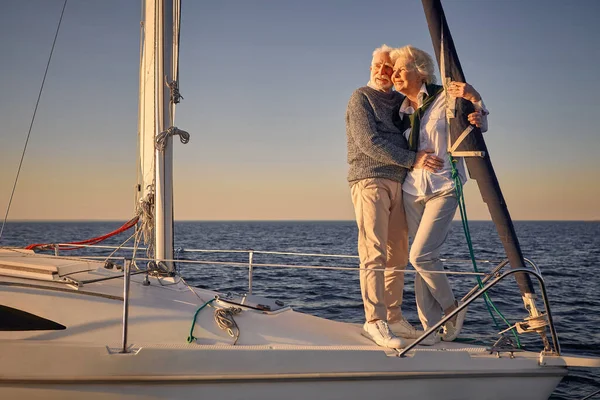 Полная длина красивой романтической пожилой пары, стоящей на борту парусника или палубы, плавающей в спокойном синем море, обнимающей и наслаждающейся удивительным закатом — стоковое фото