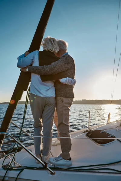 Rear view of senior couple hugging and looking at the horizon, enjoying amazing sunset while standing on the side of sail boat or yacht deck floating in sea — Stock Photo, Image