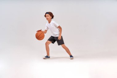 Çocuklar için en iyi spor. Gri arka planda tek başına duran, basketbol oynayan bir çocuğun tam boy çekimi.