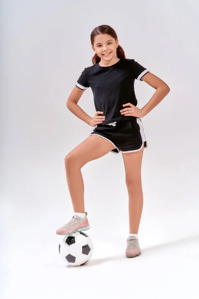 In voller Länge aufgenommen von einem niedlichen Teenager-Mädchen, das während des Trainings mit Fußball in die Kamera lächelt, isoliert vor grauem Hintergrund im Studio — Stockfoto