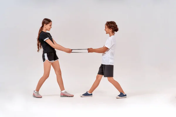 Повнометражний знімок щасливого хлопчика-підлітка і дівчинки, що розважається під час тренувань з групою опору, стоїть ізольовано на сірому фоні — стокове фото