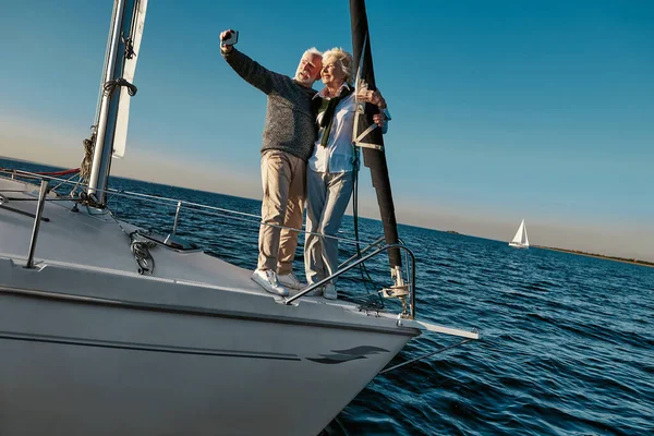 捕捉光明的时刻。年轻貌美的老夫妇站在帆船或游艇甲板上，一边在海上漂泊一边，一边用智能手机自拍 — 图库照片