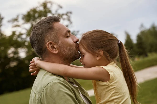 Joven padre cariñoso besando a su linda hija en la frente mientras pasan tiempo juntos al aire libre en un día de verano, sentado en una hierba verde en el parque — Foto de Stock