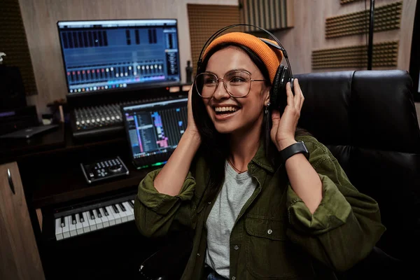 Retrato de una hermosa joven morena, artista femenina sonriendo a un lado, ajustando auriculares mientras está sentada en un estudio de grabación —  Fotos de Stock