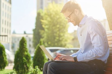 Güneşli bir günde bankta otururken dizüstü bilgisayarda çalışan yakışıklı bir erkek ya da işadamının yan görüntüsü