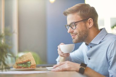 Kafede otururken kahve içip sandviç yiyen, işten önce kahvaltı yapan heyecanlı genç bir adamın yan görüntüsü.