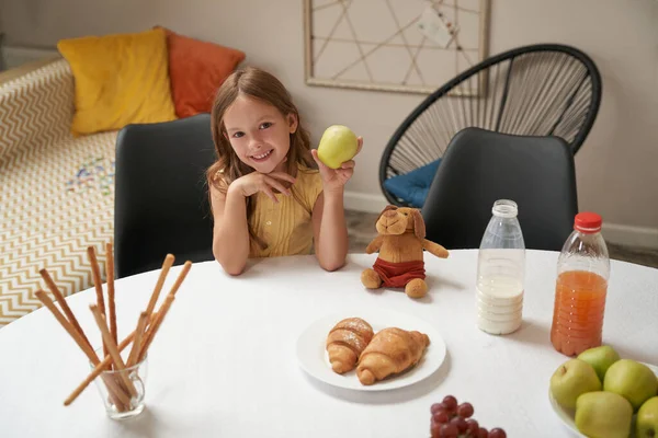 Happy caucasian girl sitting at the table at home, holding green apple and smiling at camera, having breakfast at home