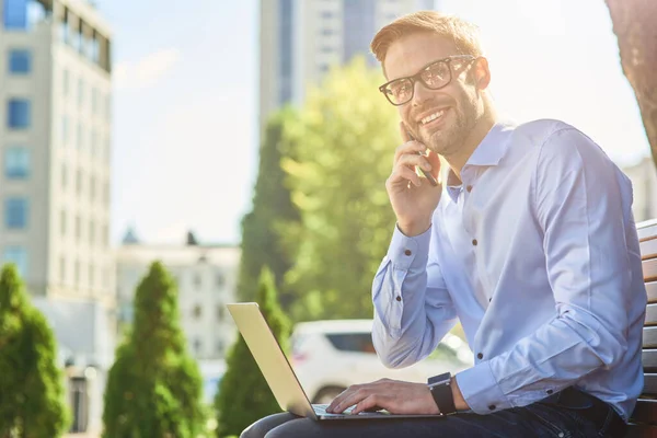 Buenas noticias. Joven hombre de negocios feliz hablando por teléfono y y sonriendo mientras está sentado en el banco al aire libre y trabajando en línea, utilizando el ordenador portátil — Foto de Stock