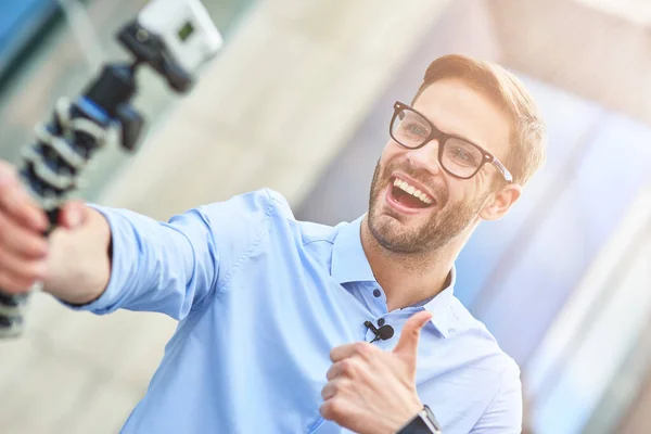 Молодой счастливый человек, мужчина-блоггер в синей рубашке, держащий подвеску со смартфоном и записывающий видео для своего блога на открытом воздухе, улыбающийся в камеру и показывающий большой палец — стоковое фото
