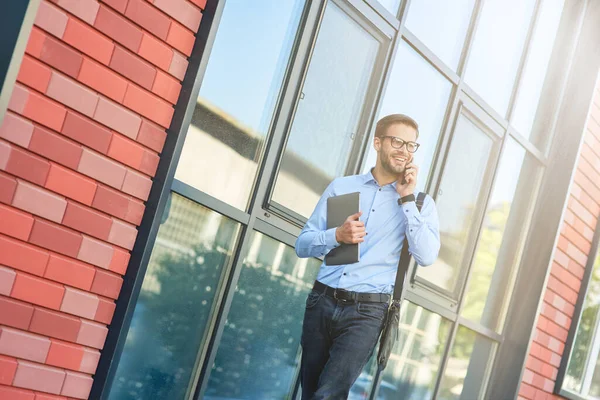 Joven hombre de negocios caucásico feliz vistiendo camisa azul y anteojos sosteniendo su computadora portátil, hablando por teléfono y sonriendo mientras camina al aire libre — Foto de Stock