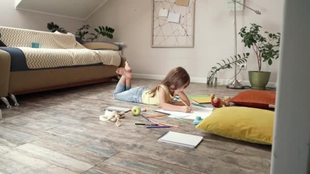 Boční pohled roztomilá holčička ležící na dřevěné podlaze v obývacím pokoji doma a kreslení s barevnými tužkami na bílém listu papíru, školka dítě hrát sám — Stock video