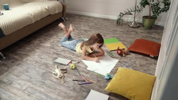 Gericht schattig klein meisje liggend op de houten vloer in de woonkamer thuis en tekening met kleurrijke potloden op een wit vel papier, het ontwikkelen van creativiteit — Stockvideo