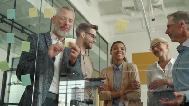Boldog multiracionális üzletemberek ragacsos jegyzeteket használnak az üvegtáblán, ötleteket osztanak meg munkatársaikkal és megvitatják a projektet, miközben az irodai tárgyalóban állnak. — Stock videók