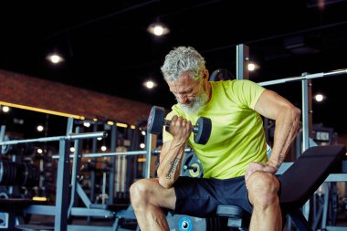 Kollarım güçleniyor. Spor kıyafetleriyle odaklanmış, ağırlık kaldıran ve spor salonunda egzersiz yaparken bisepslerini pompalayan olgun bir adam.
