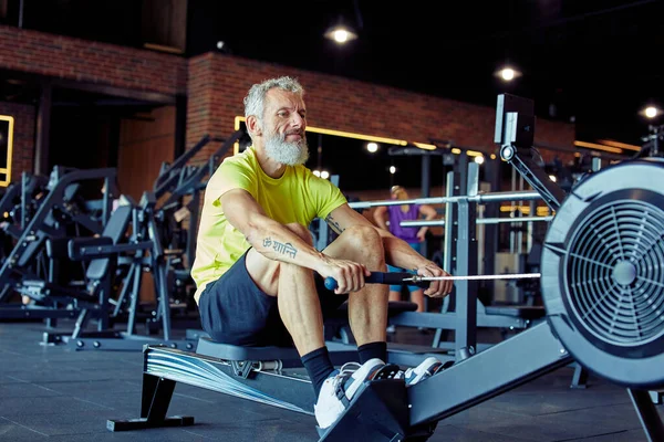 Hård träning. Gammal atletisk man i sportkläder som tränar på roddmaskin på gym — Stockfoto