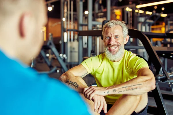 Portrait of a happy middle aged man discussing training results with fitness instructor or personal trainer and smiling while sitting together on the floor at gym